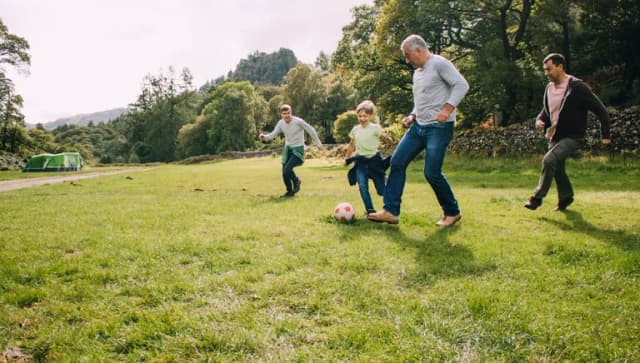 Three generation family are playing football together in a field. There are two boys, their father and their grandfather.